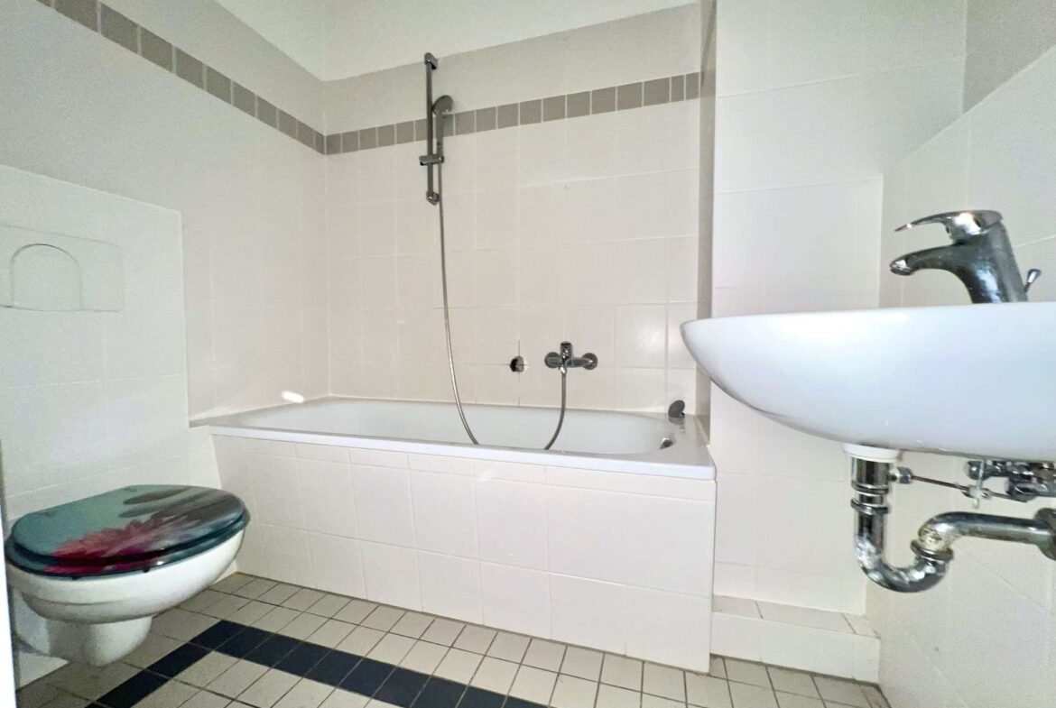 Bathroom with white tiled walls and floor, featuring a bathtub with wall-mounted shower, a toilet with a colorful seat, and a white sink with exposed chrome plumbing.