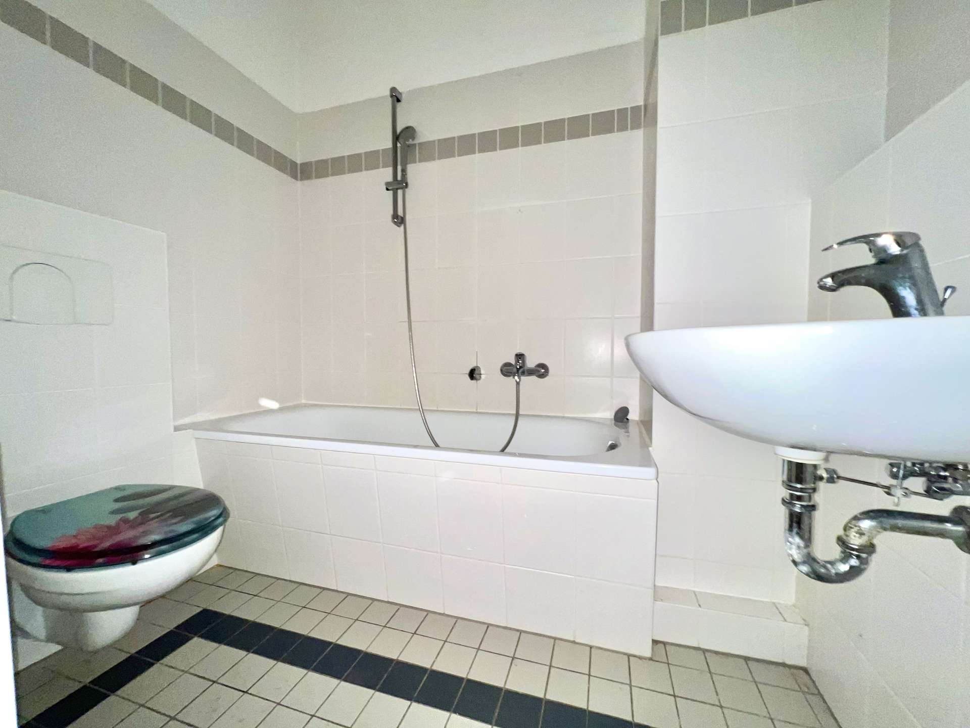 Bathroom with white tiled walls and floor, featuring a bathtub with wall-mounted shower, a toilet with a colorful seat, and a white sink with exposed chrome plumbing.