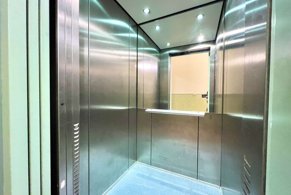 Interior of a metallic elevator cabin with stainless steel walls, blue floor, and overhead lights. An open doorway shows a beige wall beyond.