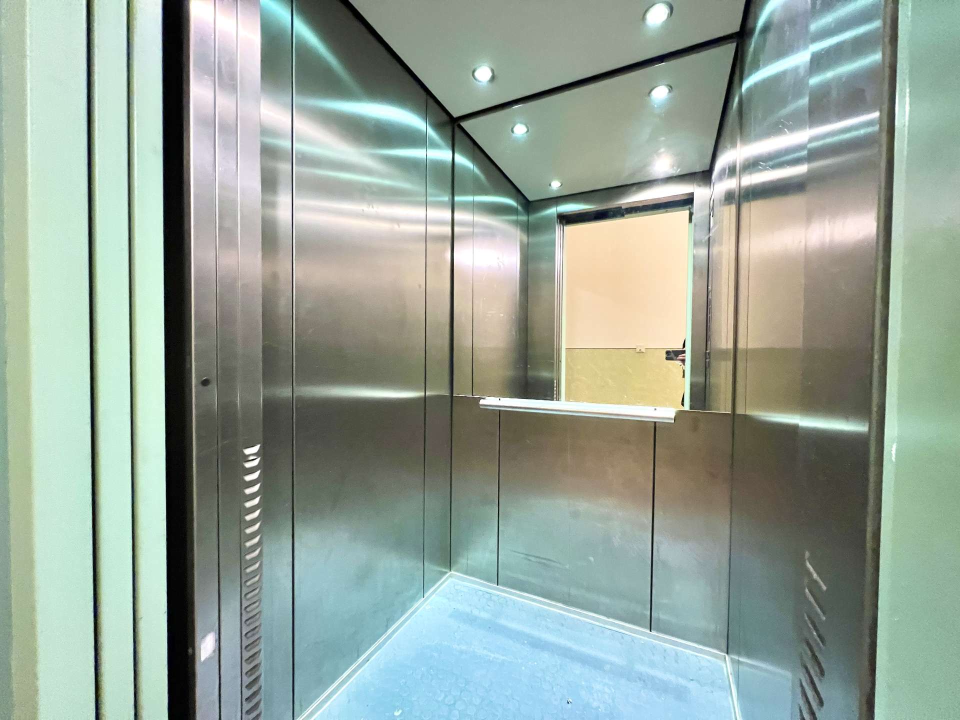 Interior of a metallic elevator cabin with stainless steel walls, blue floor, and overhead lights. An open doorway shows a beige wall beyond.