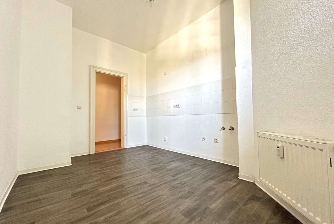 Empty living area with dark wood flooring, white walls, and a radiator on the right; doorway leads to a small tiled kitchen area.
