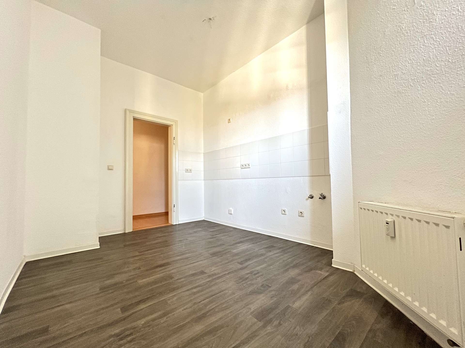 Empty living area with dark wood flooring, white walls, and a radiator on the right; doorway leads to a small tiled kitchen area.