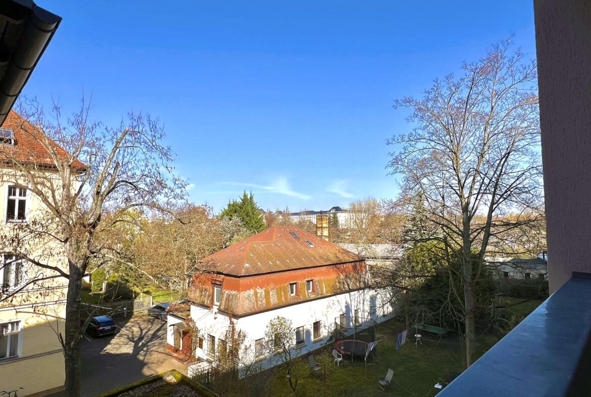 Sunny view over a courtyard with red-tiled roofs, leafless trees, and a grassy area with white chairs under a clear blue sky.