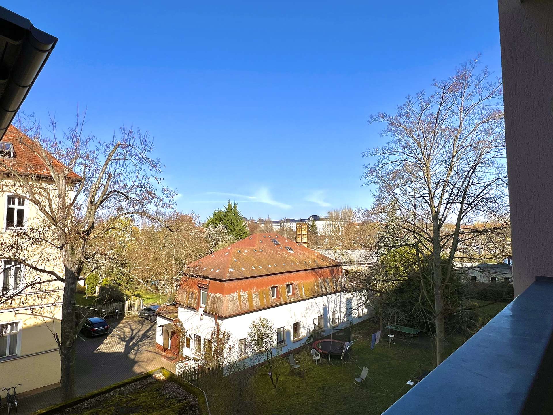 Sunny view over a courtyard with red-tiled roofs, leafless trees, and a grassy area with white chairs under a clear blue sky.