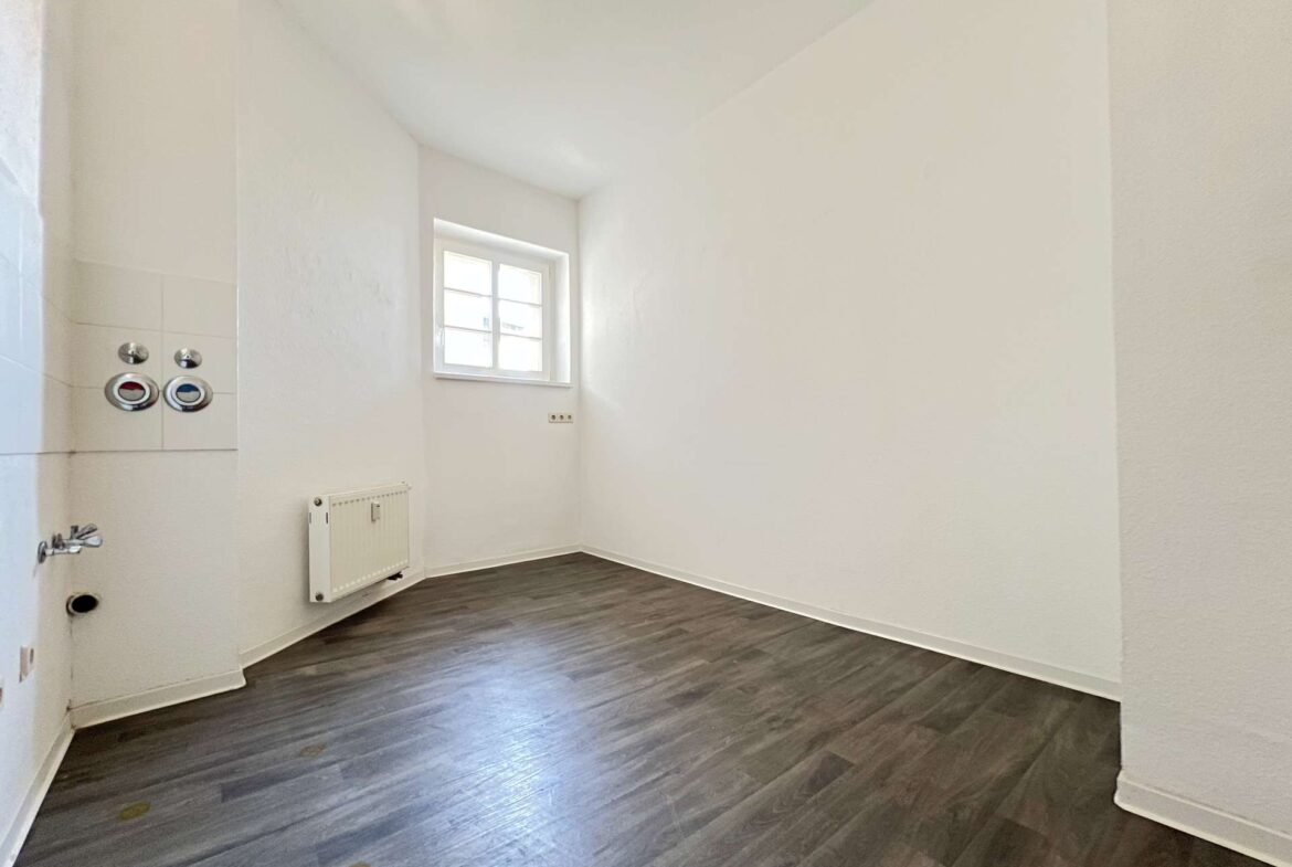 Empty white room with dark wood floor, small window, radiator, and plumbing fixtures along the left wall