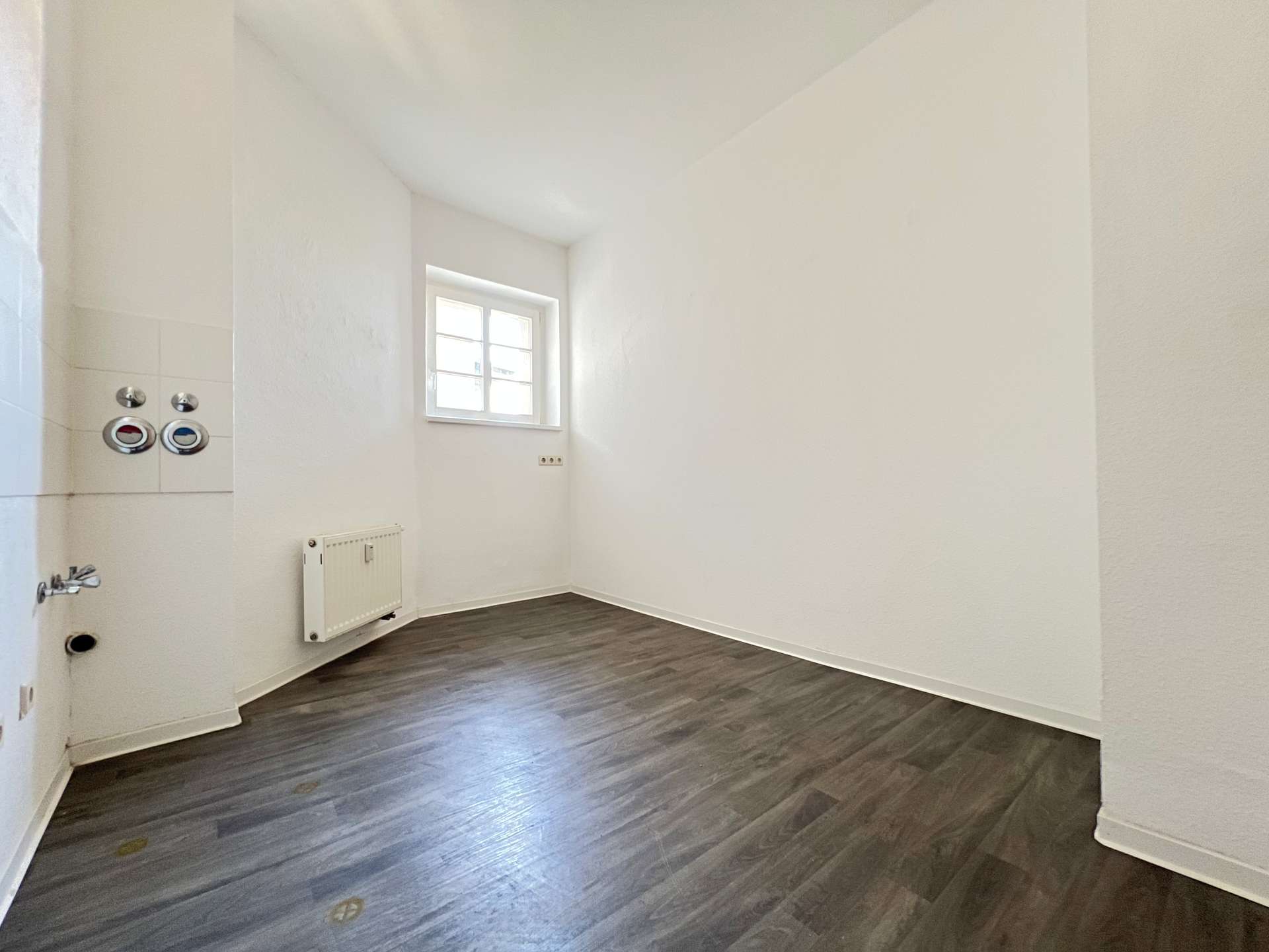 Empty white room with dark wood floor, small window, radiator, and plumbing fixtures along the left wall