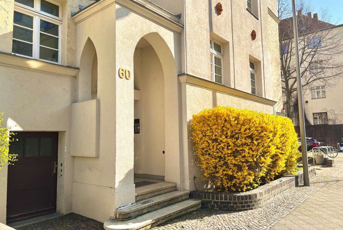 Beige apartment building entrance with an arched doorway and the number 60, a yellow shrub in front, and cobblestone path.