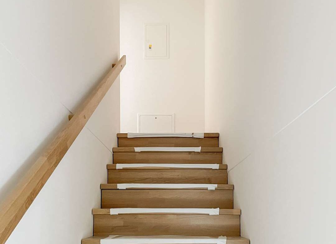 Wooden staircase under construction with taped treads and a left-hand wooden handrail; white walls and exposed wiring overhead.