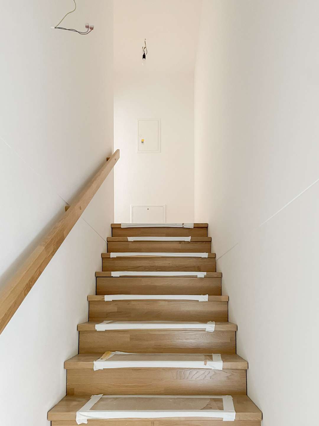 Wooden staircase under construction with taped treads and a left-hand wooden handrail; white walls and exposed wiring overhead.