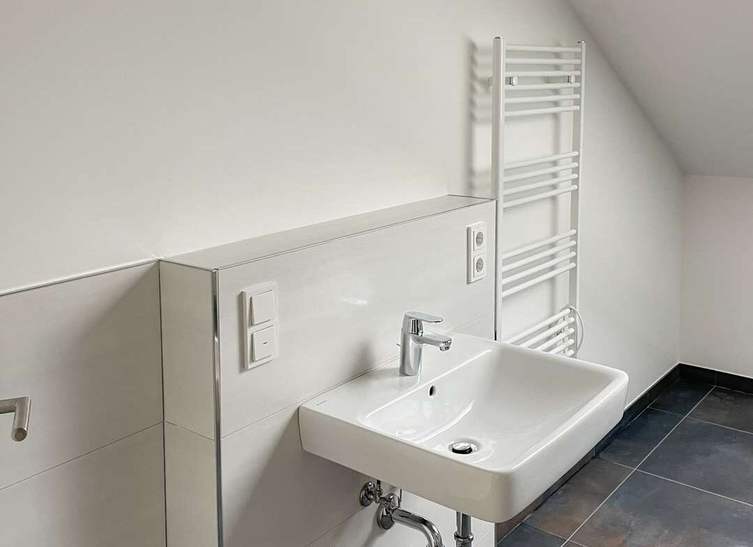 White bathroom with a wall-mounted sink, chrome faucet, and a white heated towel rail; exposed wiring above the wall.** Note: unfinished electrical work.