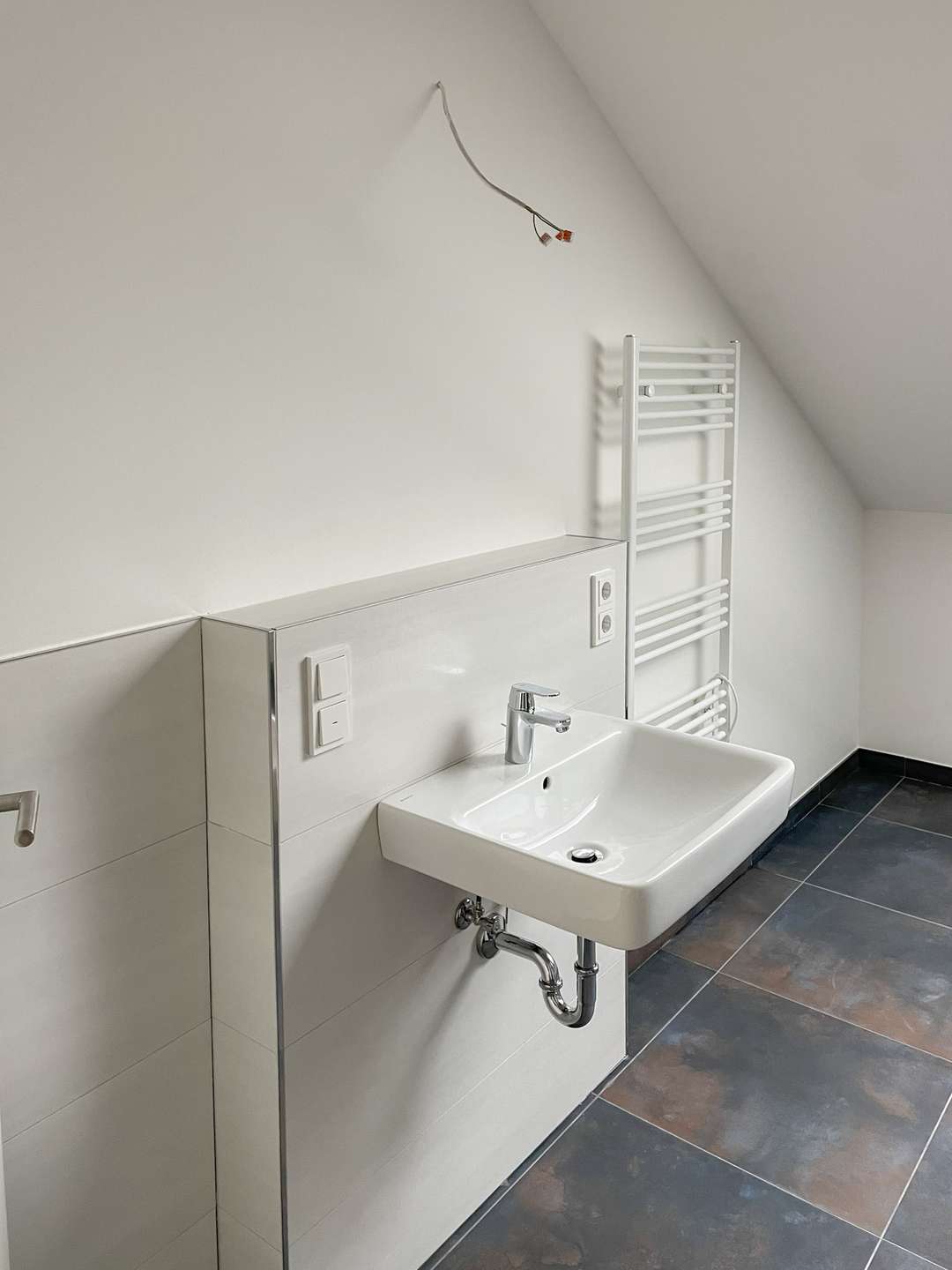 White bathroom with a wall-mounted sink, chrome faucet, and a white heated towel rail; exposed wiring above the wall.** Note: unfinished electrical work.