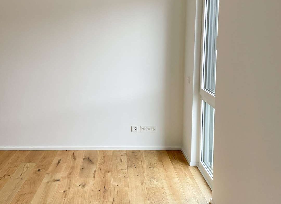 Empty white room with light wood floor, a single hanging bulb, and a large window on the right.