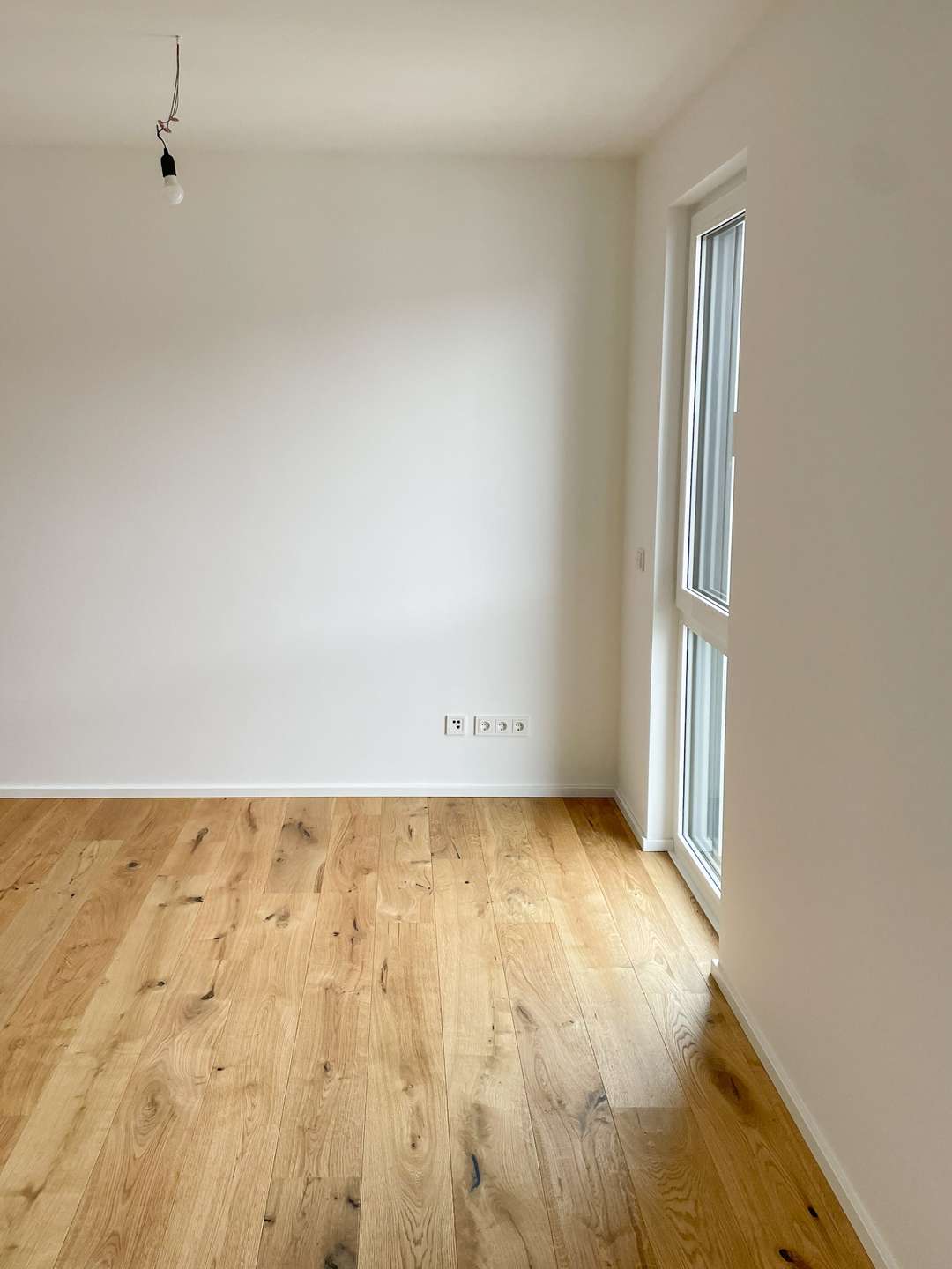 Empty white room with light wood floor, a single hanging bulb, and a large window on the right.