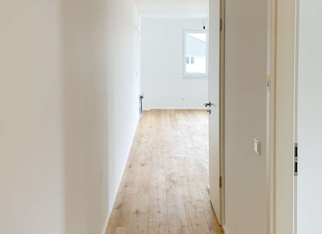 Bright empty hallway with light wood floor and white walls, open doorway to a sunlit room at the end.