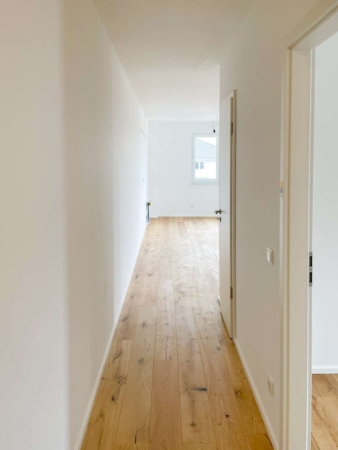 Bright empty hallway with light wood floor and white walls, open doorway to a sunlit room at the end.