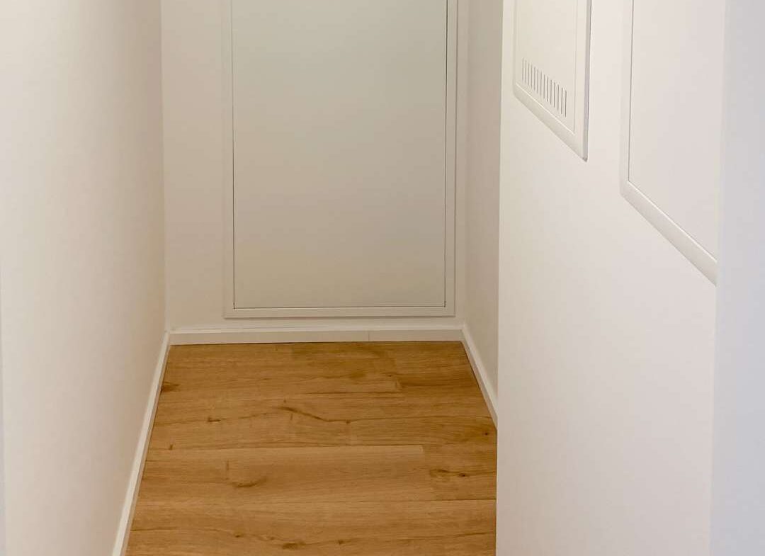 Narrow white hallway with light wood flooring and a rectangular utility access panel at the far end.