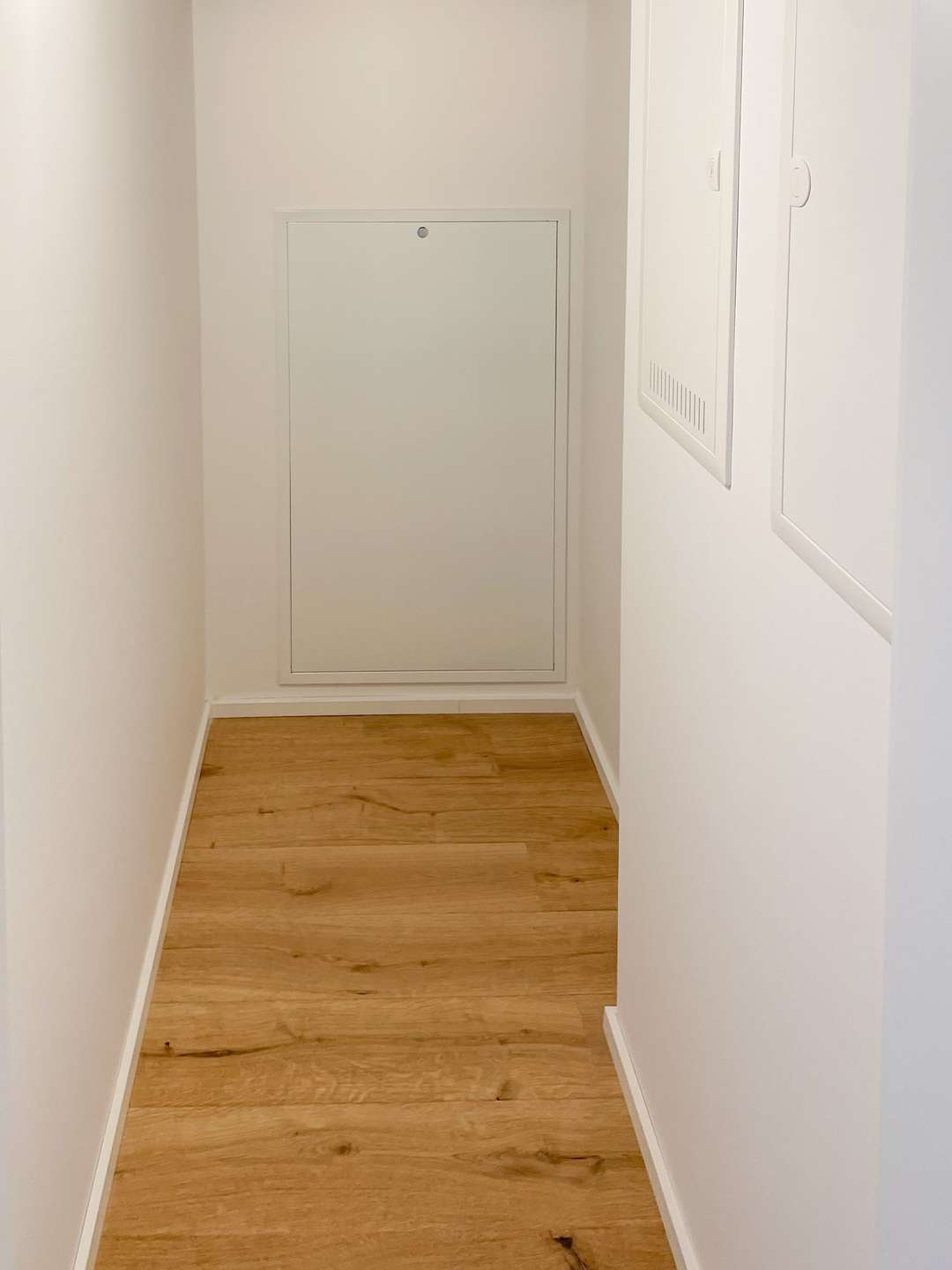 Narrow white hallway with light wood flooring and a rectangular utility access panel at the far end.