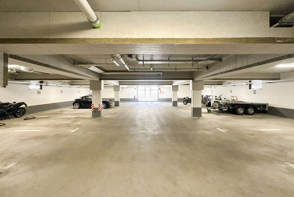 Underground parking level with concrete pillars, a few parked cars and motorcycles, and a trailer; overhead pipes and ductwork visible.