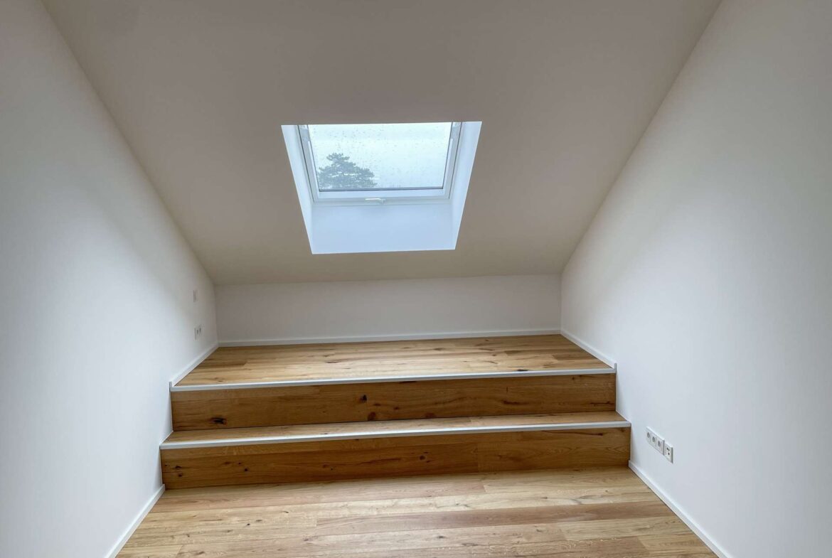 Attic room with a skylight in a sloped ceiling, white walls, and three-tier wooden steps with a built-in storage chest along the wall.