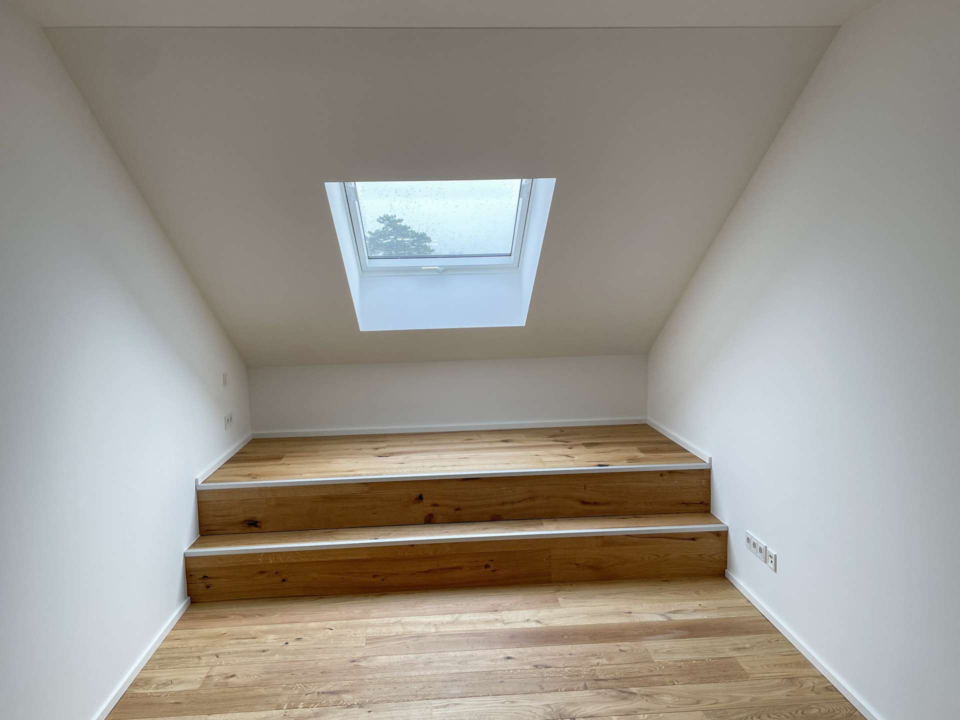 Attic room with a skylight in a sloped ceiling, white walls, and three-tier wooden steps with a built-in storage chest along the wall.