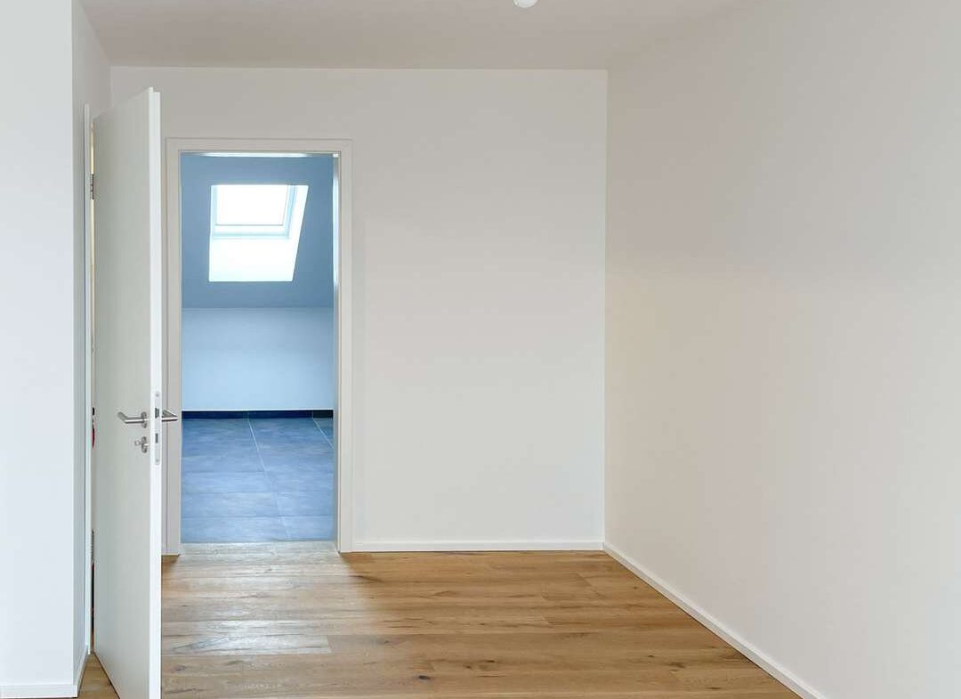 Empty white room with a wooden floor and a bare hanging light bulb; doorway opens to a blue-tiled room with a skylight afar.
