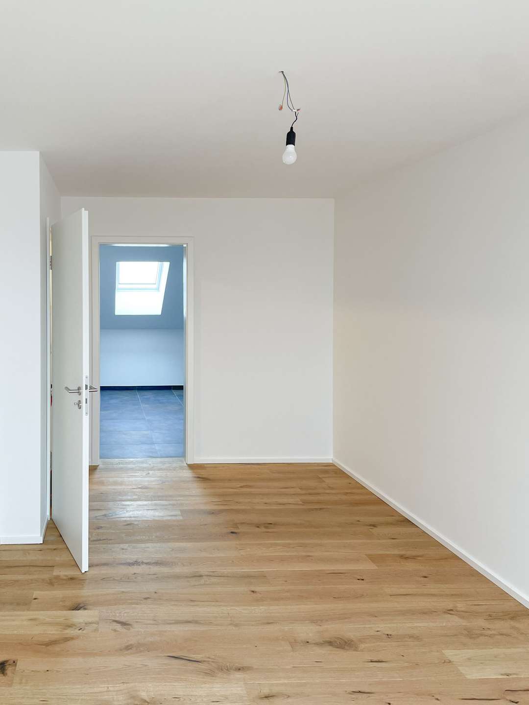 Empty white room with a wooden floor and a bare hanging light bulb; doorway opens to a blue-tiled room with a skylight afar.