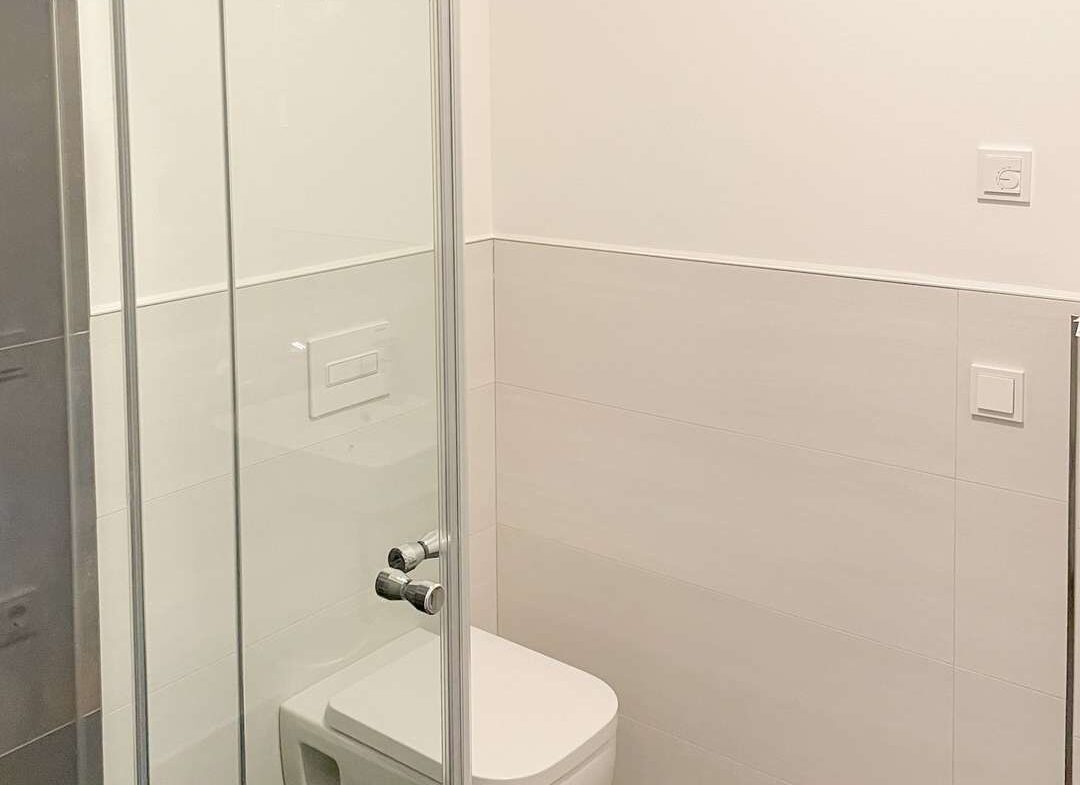 Modern bathroom with a glass shower enclosure, wall-hung toilet, and white tiled walls with dark floor tiles. Chrome fixtures add a sleek touch.