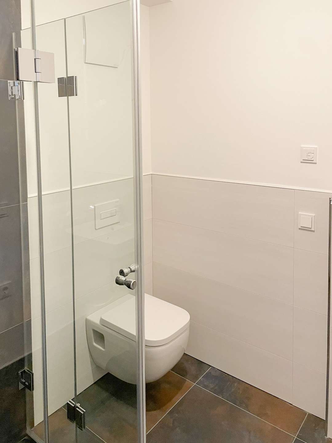 Modern bathroom with a glass shower enclosure, wall-hung toilet, and white tiled walls with dark floor tiles. Chrome fixtures add a sleek touch.