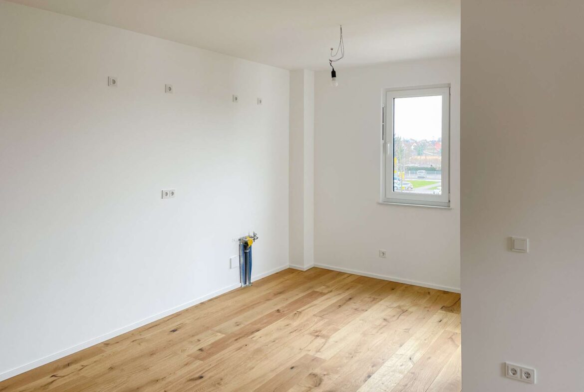 Empty white-walled room with light wood floor, a window to the outside, and a bare hanging light bulb