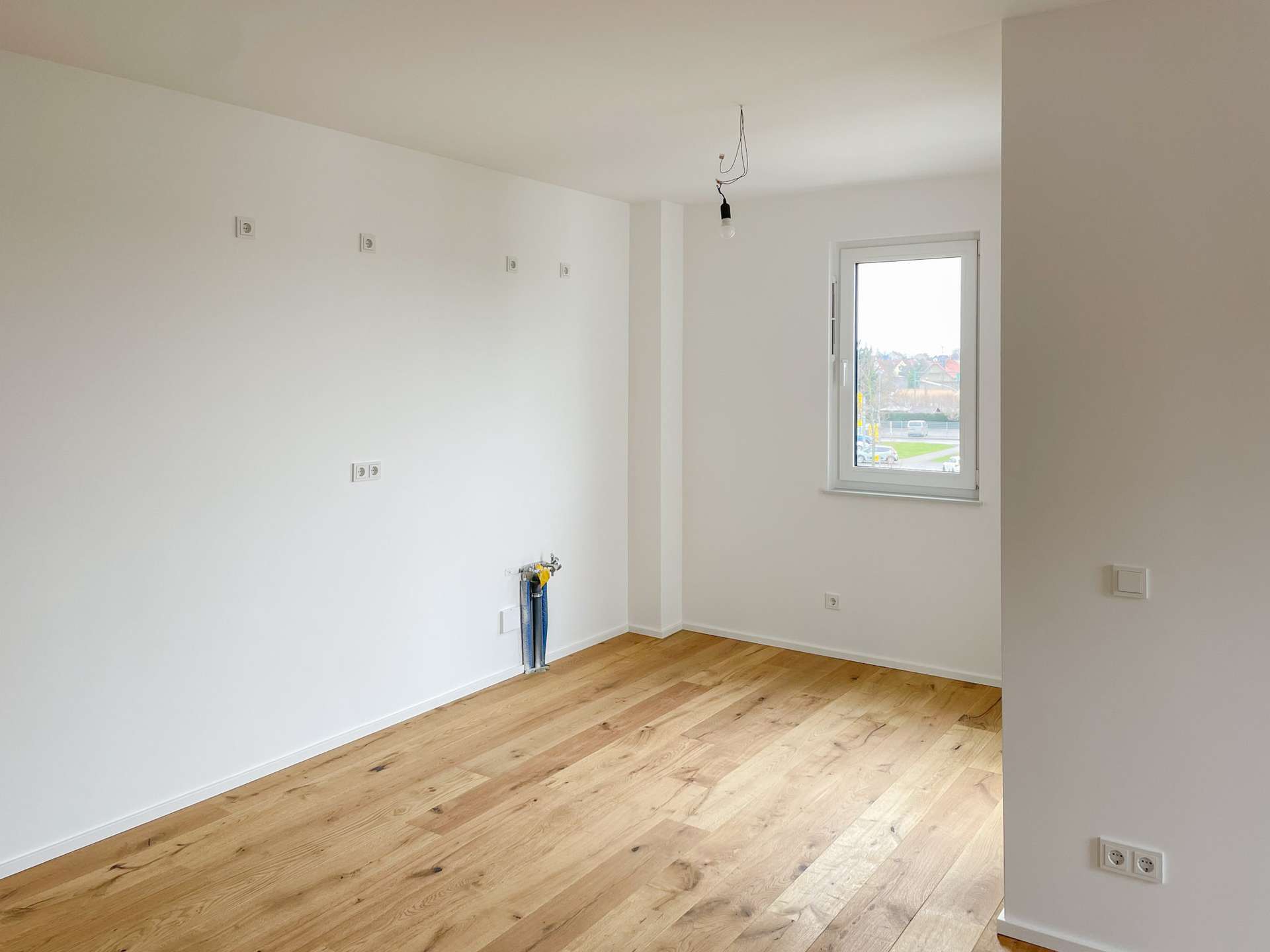 Empty white-walled room with light wood floor, a window to the outside, and a bare hanging light bulb