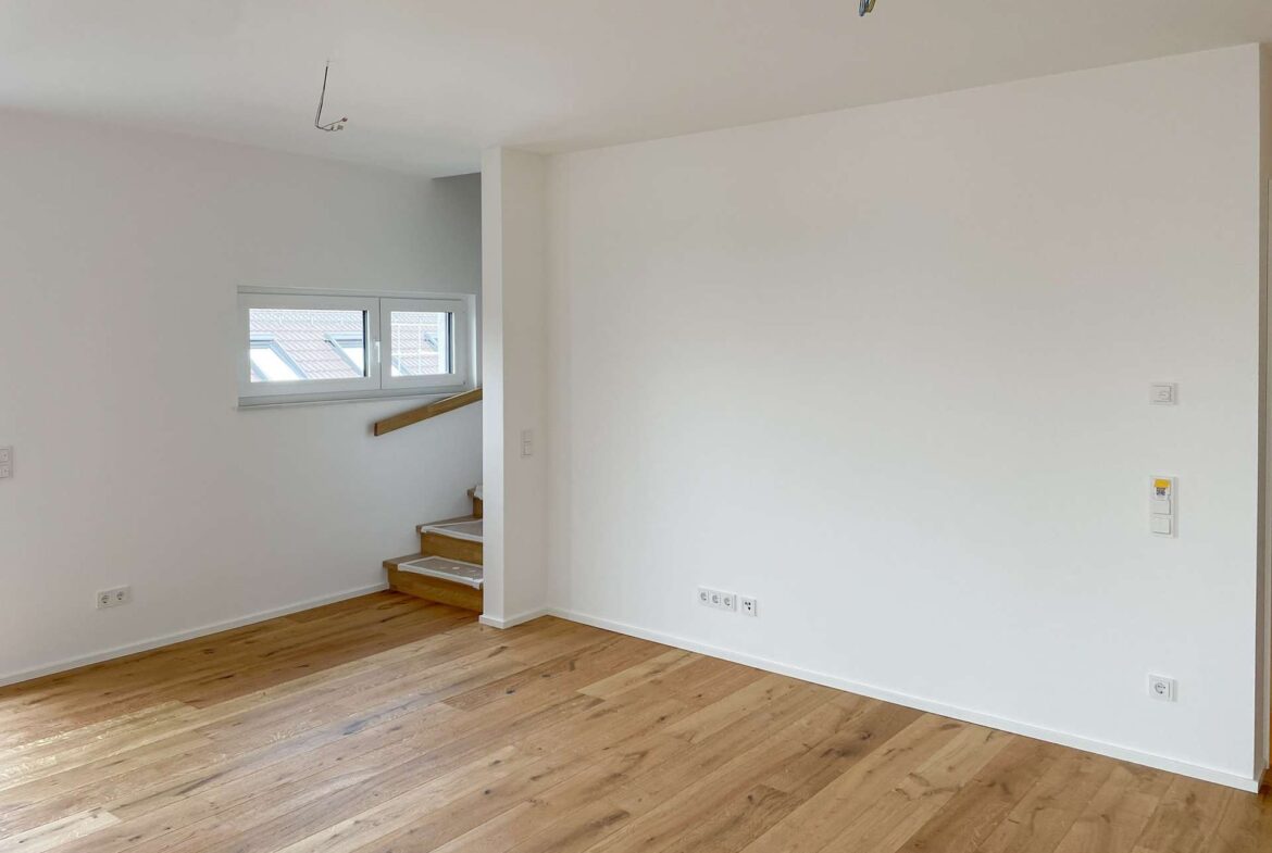 Empty living space with white walls, light wood floors, and a small staircase by a window; ceiling wires exposed.