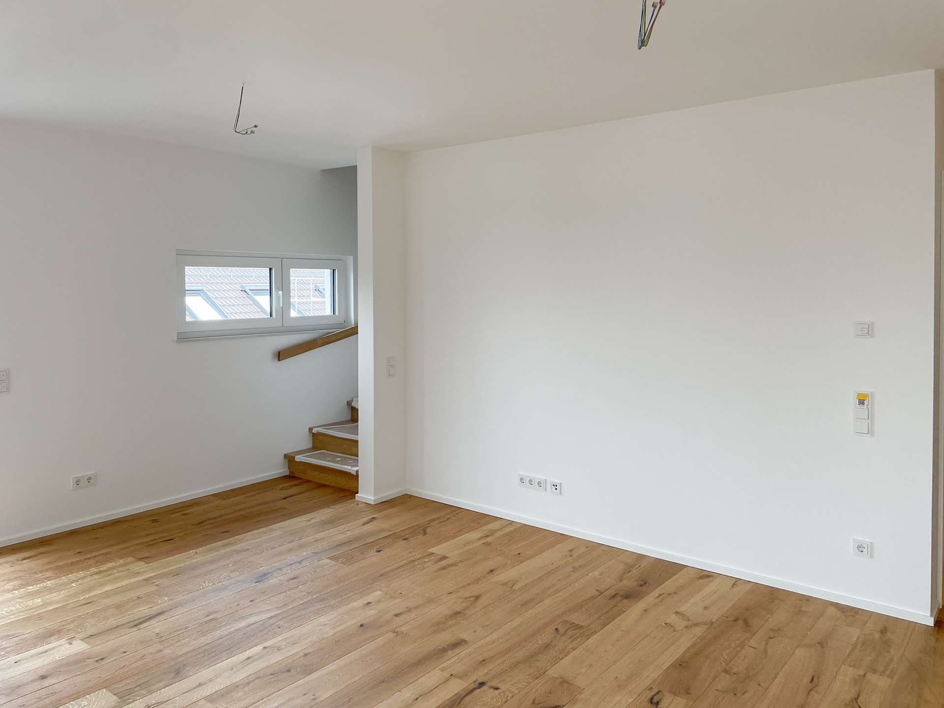Empty living space with white walls, light wood floors, and a small staircase by a window; ceiling wires exposed.