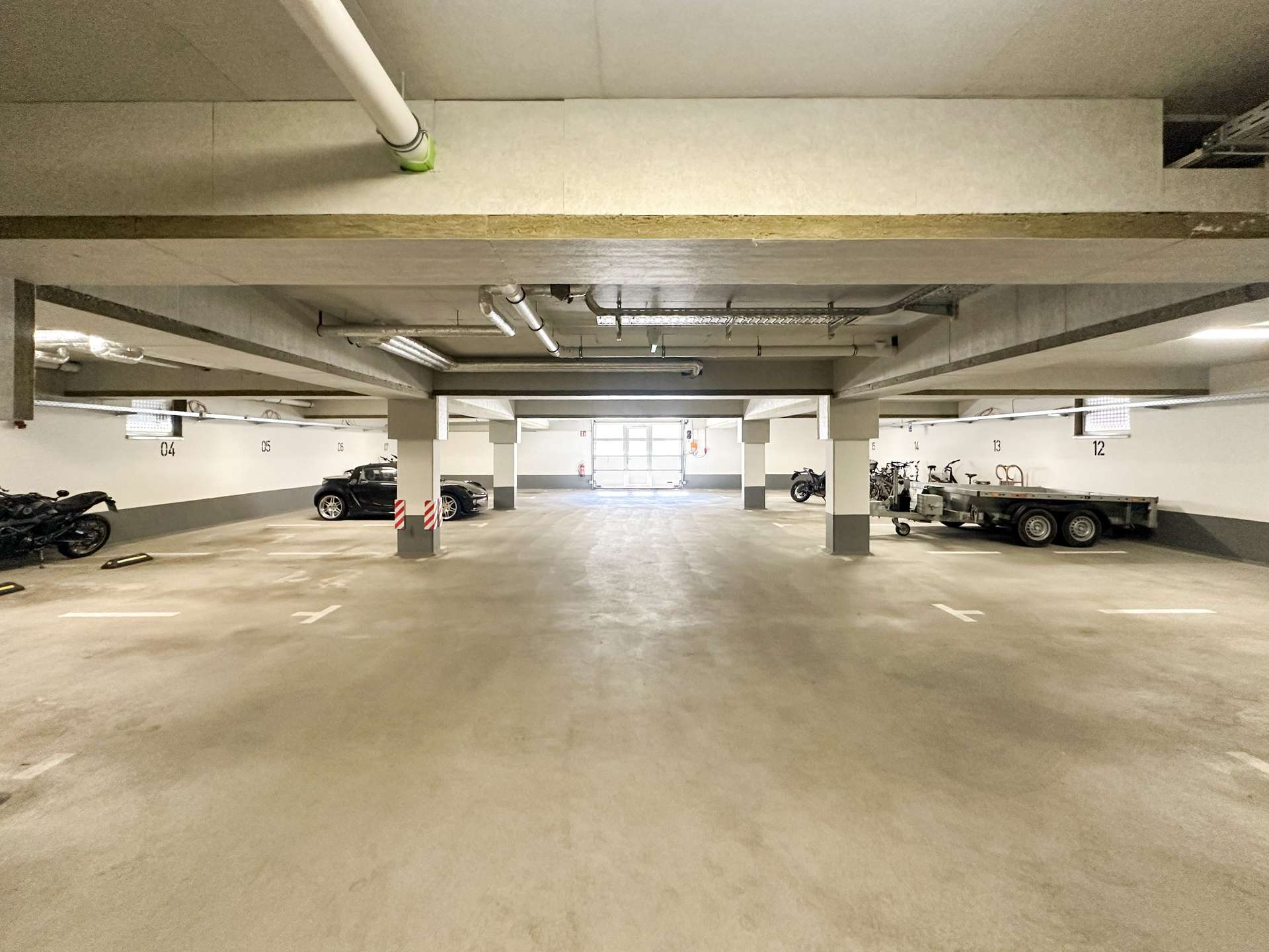 Wide view of an indoor parking garage with concrete pillars, parked cars and motorcycles along the walls, and visible ceiling pipes.