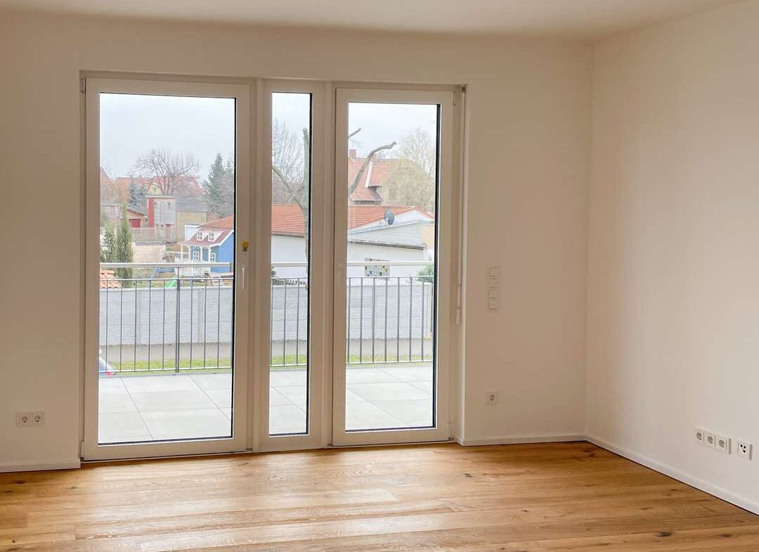 Empty living room with light wood floors and white walls, large glass doors opening to a balcony with a suburban street view outside, bare ceiling light hanging fixture above.
