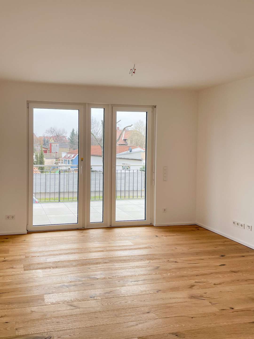Empty living room with light wood floors and white walls, large glass doors opening to a balcony with a suburban street view outside, bare ceiling light hanging fixture above.