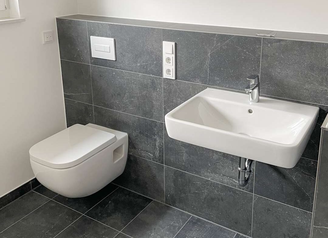 Modern bathroom with a wall-hung toilet and a white rectangular sink, set against dark gray tile walls and floor.