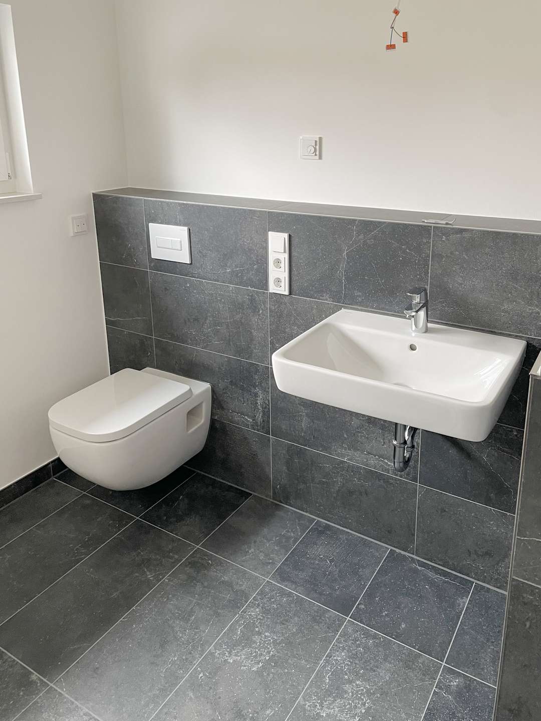 Modern bathroom with a wall-hung toilet and a white rectangular sink, set against dark gray tile walls and floor.