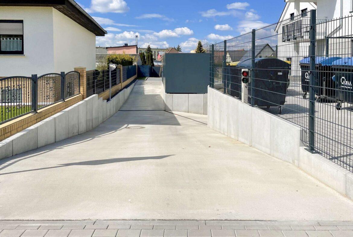 Wide concrete driveway flanked by metal fencing and low walls, leading past houses under a blue, sunny sky, with clouds above.