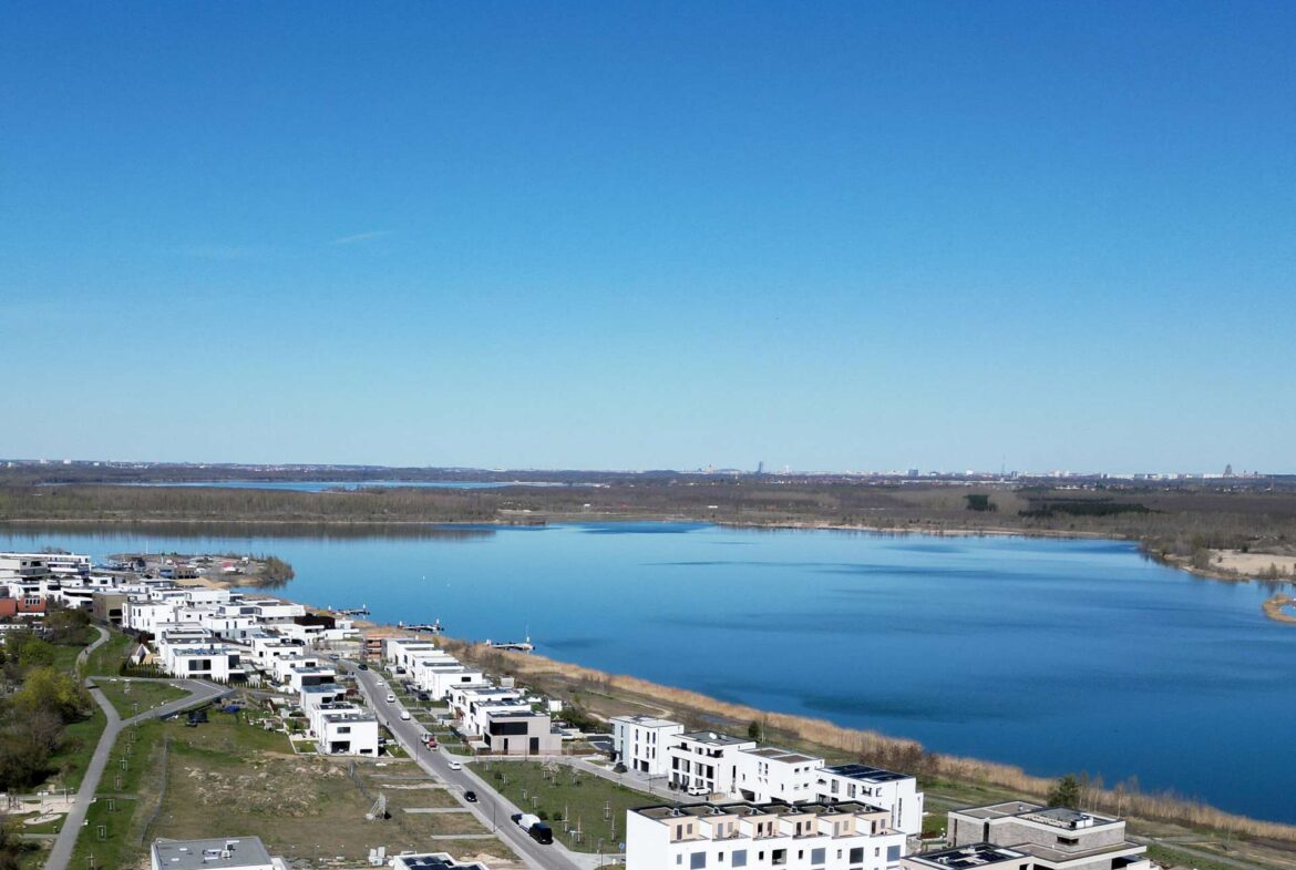 Row of modern white townhomes along a calm river under a clear blue sky, aerial view.