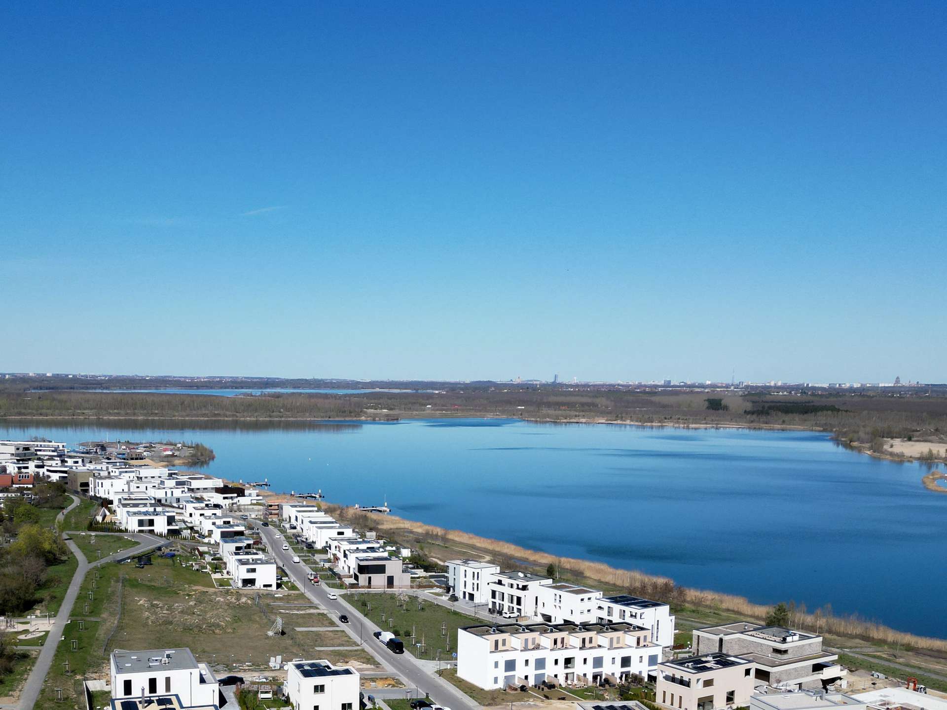 Row of modern white townhomes along a calm river under a clear blue sky, aerial view.
