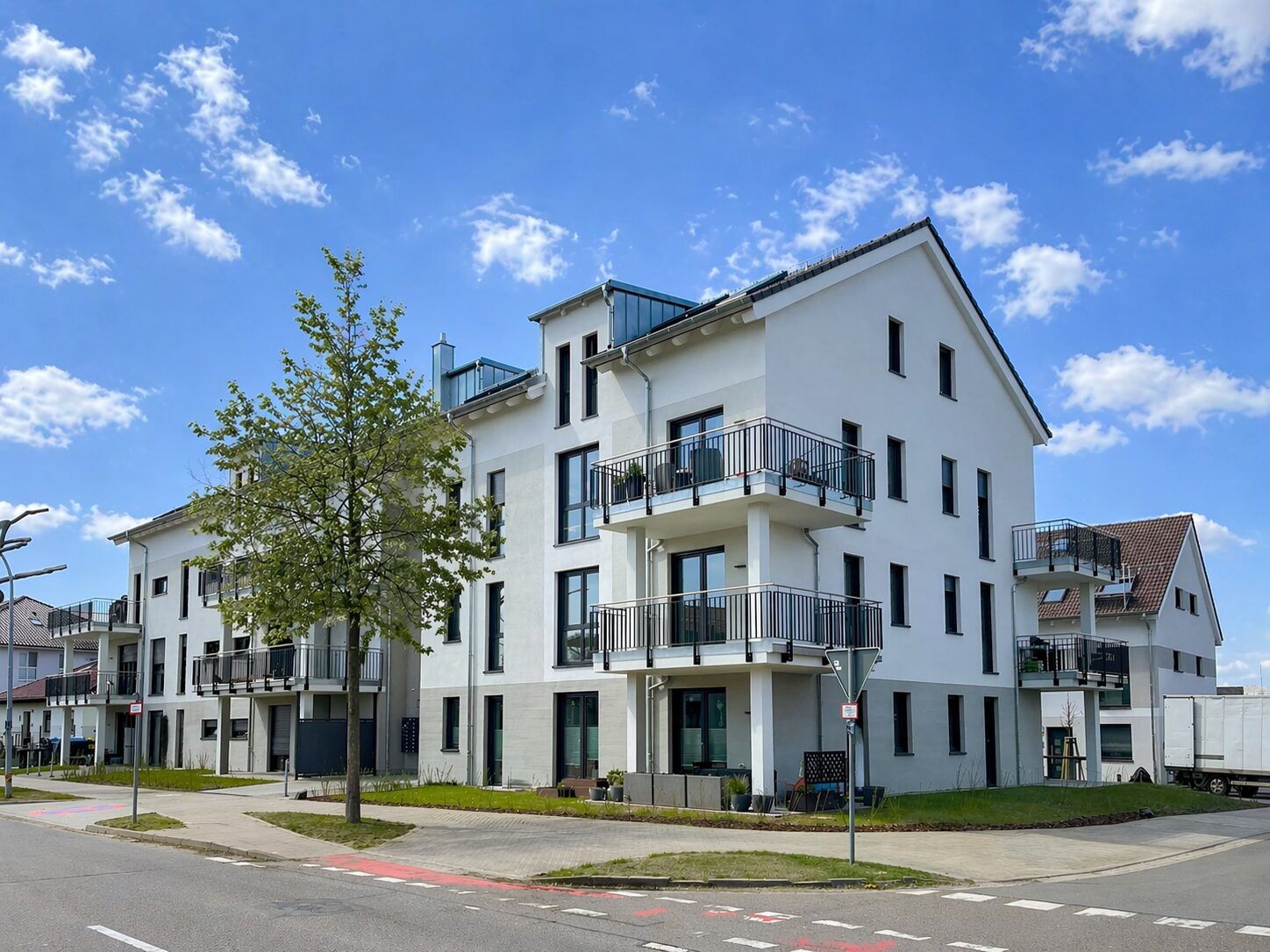 White modern apartment building with multiple balconies along a corner lot, blue sky above.
