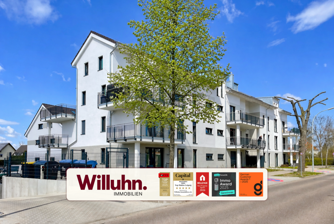 White modern apartment building with several balconies and a large tree in front, under a bright blue sky.