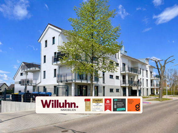 White modern apartment building with several balconies and a large tree in front, under a bright blue sky.