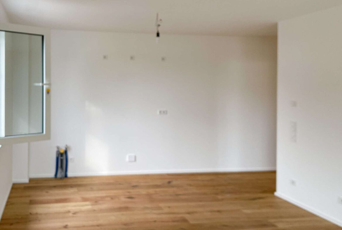 Empty white-walled room with a wooden floor, a window on the left, and a single hanging light bulb from the ceiling