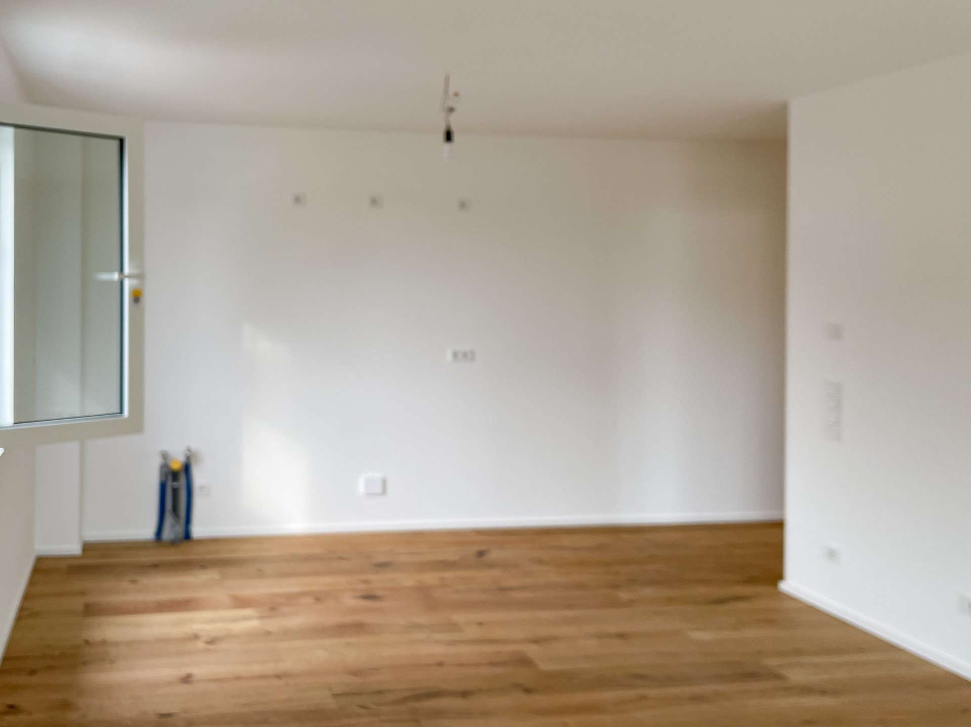 Empty white-walled room with a wooden floor, a window on the left, and a single hanging light bulb from the ceiling