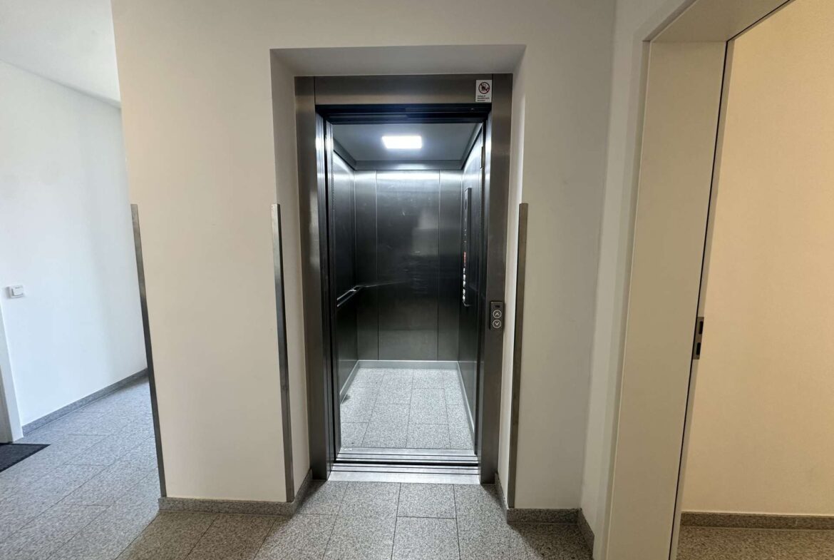 Open elevator with metal interior visible, doors partially open in a light hallway.