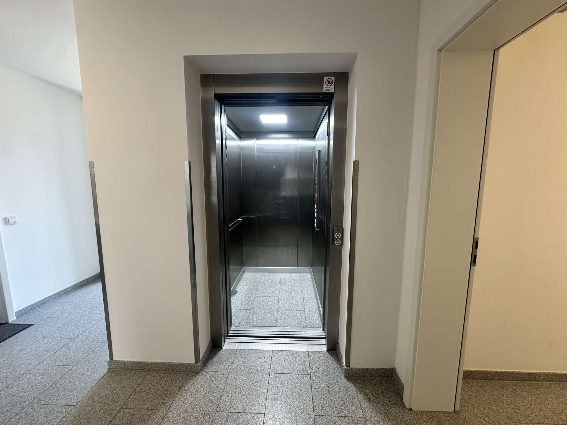 Open elevator with metal interior visible, doors partially open in a light hallway.
