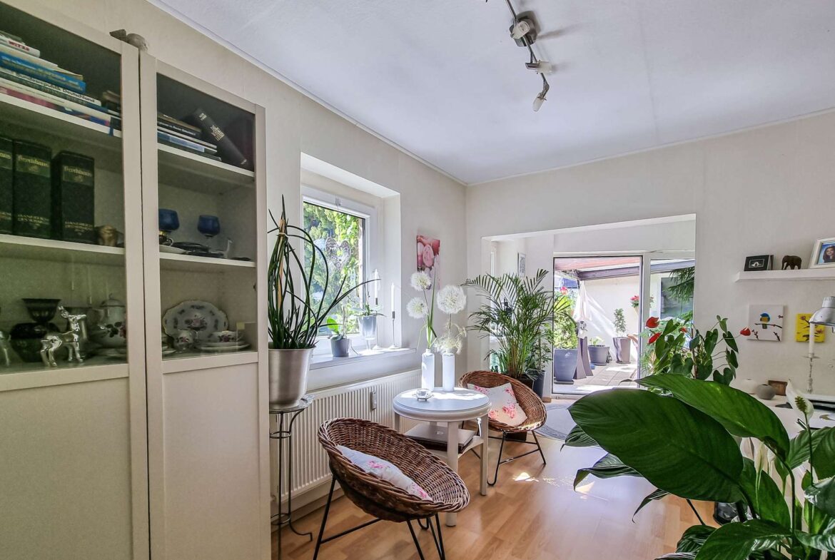 Bright living area with white display cabinets, many potted plants by the window, and wicker chairs around a small white table.