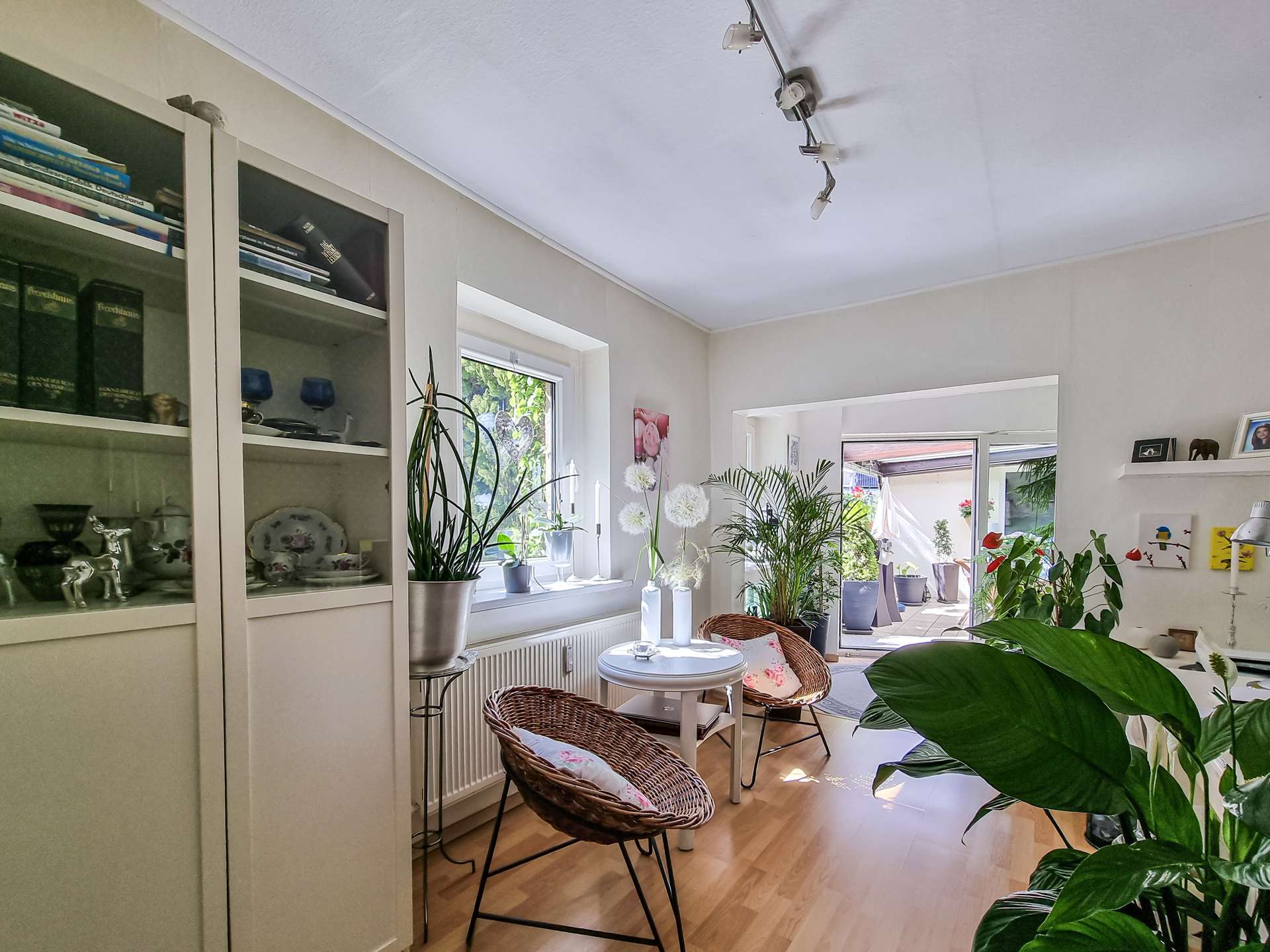 Bright living area with white display cabinets, many potted plants by the window, and wicker chairs around a small white table.