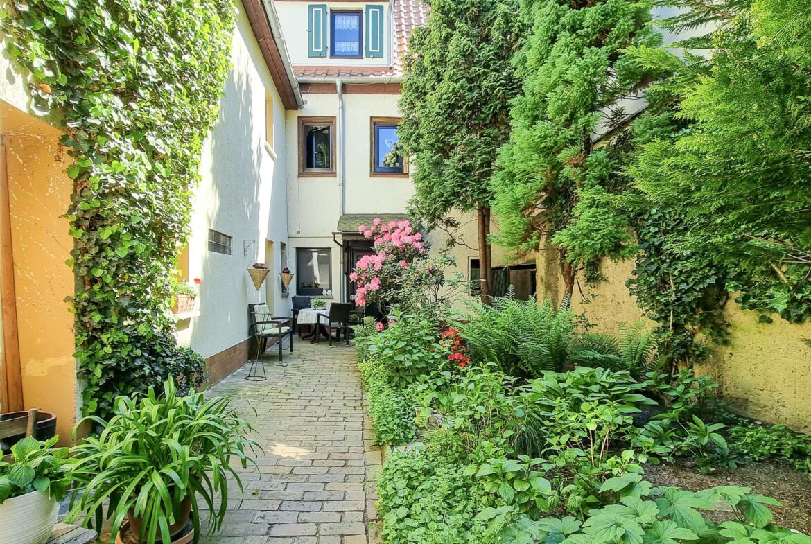 Sunlit narrow courtyard with ivy-covered walls, potted plants, and a small dining table with chairs along a brick path.